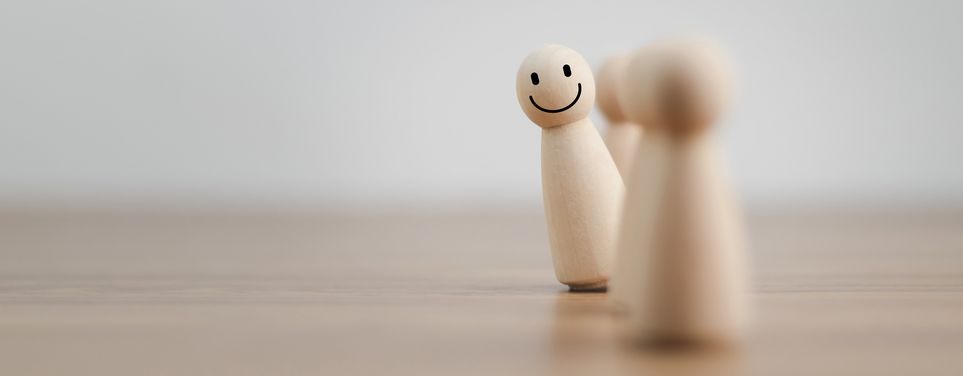 Zwei Spielfiguren aus Holz auf einer Holzunterlage. Eine Figur hat ein lächelndes Gesicht aufgemalt